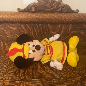 May include: A plush Mickey Mouse toy wearing a yellow and red drum major uniform. The toy is lying on a wooden surface.