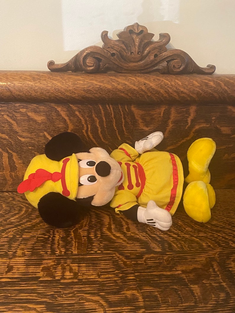 May include: A plush Mickey Mouse toy wearing a yellow and red drum major uniform. The toy is lying on a wooden surface.