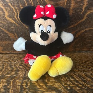 Pu&ograve; includere: Una bambola di peluche di Minnie Mouse con pelliccia nera, un fiocco rosso a pois bianchi e piedi gialli. La bambola ha guanti bianchi e una gonna rossa e bianca a pois. La bambola &egrave; seduta su una superficie di legno.
