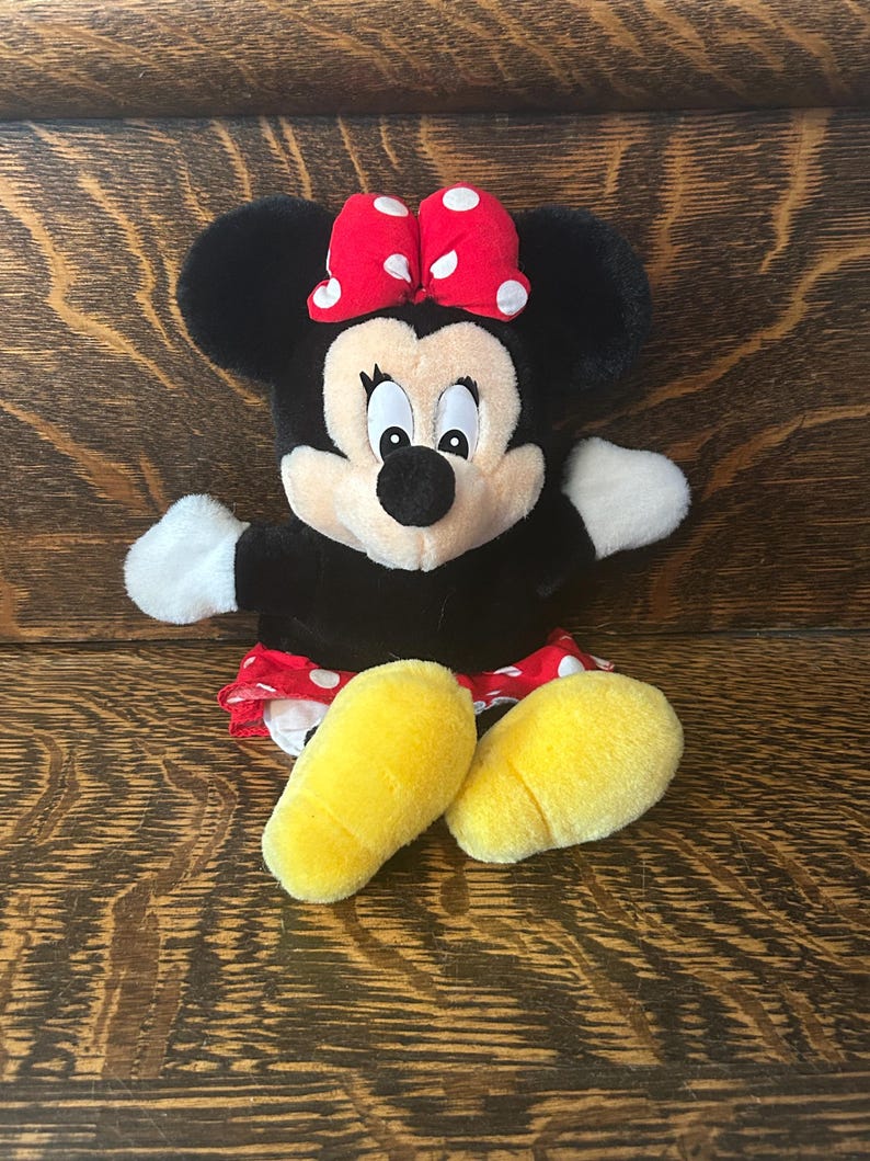 Pu&ograve; includere: Una bambola di peluche di Minnie Mouse con pelliccia nera, un fiocco rosso a pois bianchi e piedi gialli. La bambola ha guanti bianchi e una gonna rossa e bianca a pois. La bambola &egrave; seduta su una superficie di legno.