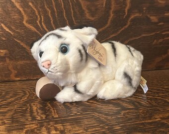 May include: White plush toy tiger cub with black stripes, blue eyes, and a pink nose. The tiger is lying down with its head turned to the side and its paw resting on a brown ball. The toy has a tag that says "Myoni's Totos".