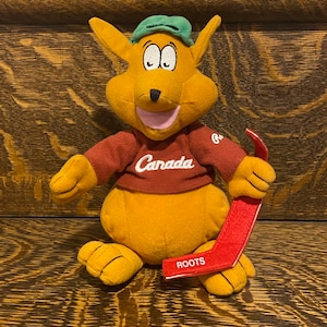 May include: A plush brown kangaroo wearing a red shirt with "Canada" written on it and a green hat. The kangaroo is holding a red hockey stick with "ROOTS" written on it.