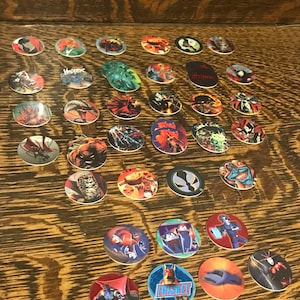 May include: A collection of oval-shaped collectible game pieces, featuring various comic book characters and designs. The pieces are arranged on a wooden surface, showcasing a variety of colors and illustrations. Some pieces have text, including the word "Frisket".
