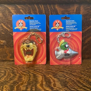 May include: Two Looney Tunes keychain clocks. The clock on the left features Taz the Tasmanian Devil with a red and yellow design. The clock on the right features Marvin the Martian in a silver rocket ship. Both clocks have a digital display showing 12:08.