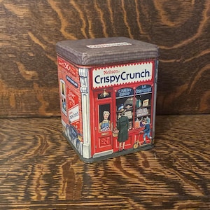 Può includere: Una scatola di latta vintage con un'illustrazione rossa, bianca e blu di un negozio di dolciumi. Il testo "Neilson Crispy Crunch" è stampato sulla parte anteriore della scatola.