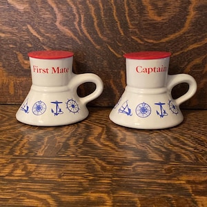 May include: Two white ceramic mugs with red lids and blue nautical designs. The mugs are labeled "First Mate" and "Captain".