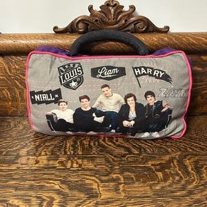 May include: Gray pillow with a black handle and a pink border. The pillow features images of five people and the names "Louis", "Liam", "Harry", "Zayn", and "Niall". The pillow is on a wooden surface.