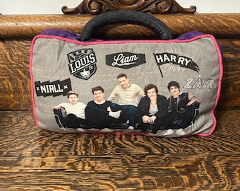 May include: Gray pillow with a black handle and a pink border. The pillow features images of five people and the names "Louis", "Liam", "Harry", "Zayn", and "Niall". The pillow is on a wooden surface.