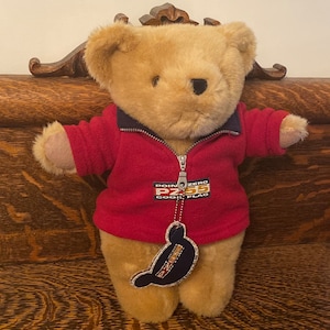May include: A brown teddy bear wearing a red fleece zip-up jacket with a blue collar and the text "Point Zero P255 Code Flag". The bear is holding a blue and silver tag with a Mickey Mouse silhouette.
