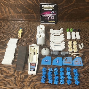 May include: A vintage Construx Stellar Exploration Set by Fisher-Price including an instruction manual, a small astronaut figure, various white structural beams, blue wing panels with decals, grey connectors, and small blue building blocks.