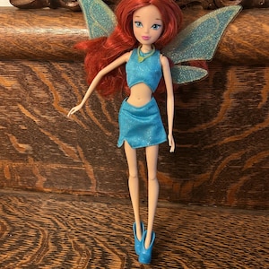 Puede incluir: Muñeca de hada con pelo rojo, ojos azules y un atuendo turquesa. La muñeca tiene alas brillantes y lleva una corona y zapatos azules. La muñeca está sobre una superficie de madera.