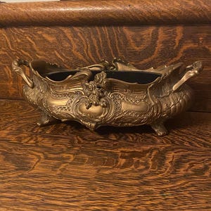 May include: A vintage brass planter with ornate details and a black interior. The planter has a rounded oval shape with two handles on either side. The planter is sitting on a wooden surface.