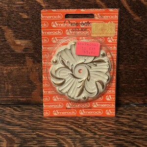 Puede incluir: Roseta decorativa para muebles Amerock en tonos marfil y dorado. La roseta está en un embalaje de plástico transparente con el logotipo de Amerock e información del producto. El embalaje está sobre una superficie de madera.