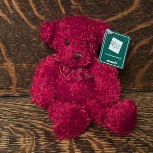May include: A plush teddy bear with deep red, textured fur, dark eyes, and a small dark nose. A shiny red ribbon is tied around its neck. A green rectangular tag is attached to the ear, featuring the text RUSS and amram's in white lettering.