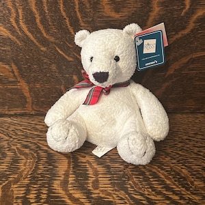 May include: A white teddy bear with a red and green tartan ribbon. The bear has a dark brown nose and small black eyes. A tag from "Ross" is attached to the bear.