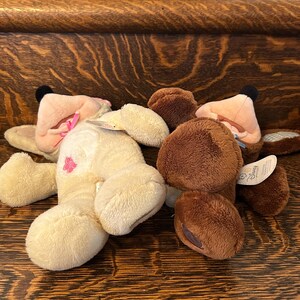 May include: Two plush stuffed animals, one brown and one cream colored, lying on a wooden surface. The cream colored plush animal has a pink bow and a pink flower on its chest. The brown plush animal has a brown nose and a brown tag that says "Disney".