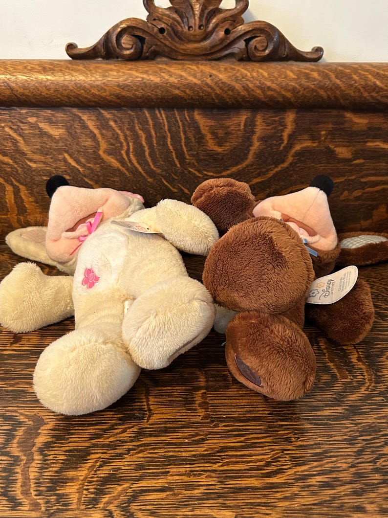 May include: Two plush stuffed animals, one brown and one cream colored, lying on a wooden surface. The cream colored plush animal has a pink bow and a pink flower on its chest. The brown plush animal has a brown nose and a brown tag that says "Disney".