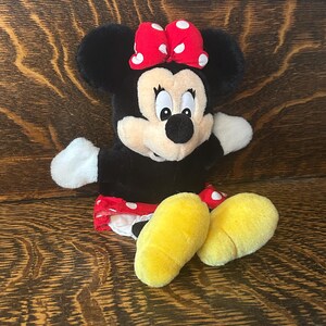 Pu&ograve; includere: Un peluche di Minnie Mouse con corpo nero, guanti bianchi e piedi gialli. Indossa un fiocco rosso a pois bianchi e una gonna rossa a pois bianchi. Il peluche &egrave; su una superficie di legno.