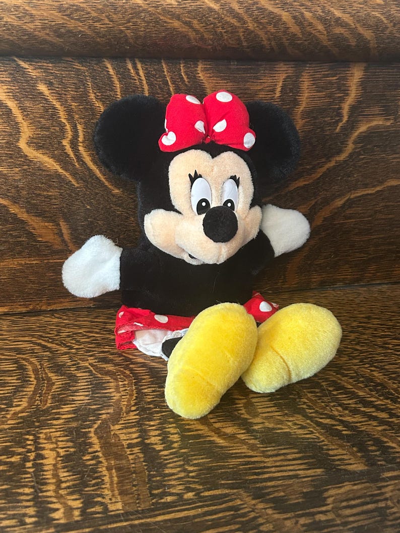 Pu&ograve; includere: Un peluche di Minnie Mouse con corpo nero, guanti bianchi e piedi gialli. Indossa un fiocco rosso a pois bianchi e una gonna rossa a pois bianchi. Il peluche &egrave; su una superficie di legno.