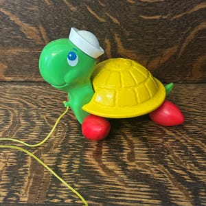 Op de afbeelding: Een vintage trek-speelgoed schildpad. De schildpad heeft een groene kop en lichaam, een gele schild en rode voeten. Het draagt een witte matrozenhoed en heeft een geel trekkoord. Het speelgoed is gemaakt van plastic en is ontworpen voor jonge kinderen.