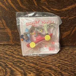 May include: A plastic bag containing a red toy car with three Looney Tunes characters. The characters are Wile E. Coyote, Road Runner, and Tweety Bird. The bag has a white card with the words "Looney Tunes" and "Wile E. Coyote" printed on it.