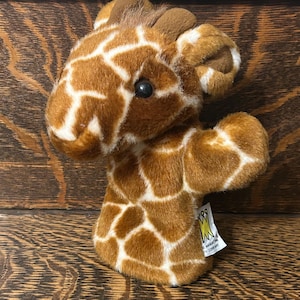 May include: A plush giraffe hand puppet with brown and white patterned fur. The puppet has a detailed face with a black eye and small ears. A tag with text is attached to the puppet.