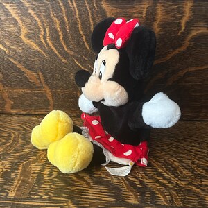 Pu&ograve; includere: Una bambola di peluche di Minnie Mouse con pelliccia nera, guanti bianchi e un fiocco rosso a pois. Indossa una gonna rossa a pois bianchi e scarpe di peluche gialle. La bambola &egrave; su una superficie di legno.