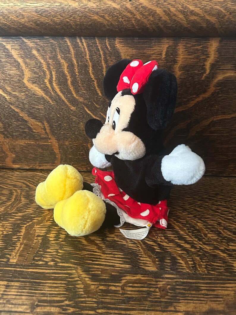 Pu&ograve; includere: Una bambola di peluche di Minnie Mouse con pelliccia nera, guanti bianchi e un fiocco rosso a pois. Indossa una gonna rossa a pois bianchi e scarpe di peluche gialle. La bambola &egrave; su una superficie di legno.