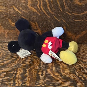 May include: A black and red plush Mickey Mouse toy with yellow shoes. The toy is lying on a wooden surface. The toy has a tag that says "Mickey Mouse" and "Disney".