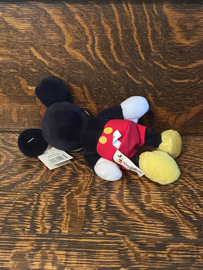 May include: A black and red plush Mickey Mouse toy with yellow shoes. The toy is lying on a wooden surface. The toy has a tag that says "Mickey Mouse" and "Disney".