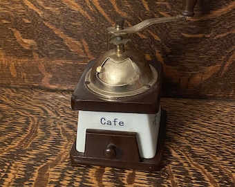 May include: A vintage wooden coffee grinder with a metal top and a brown wooden handle. The grinder has a white porcelain base with the word "Cafe" written on it.