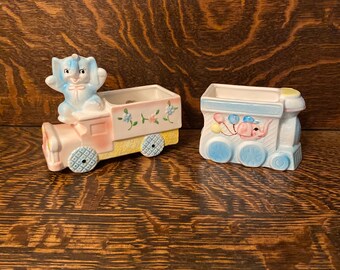 May include: Two vintage ceramic planters shaped like a train and a truck. The truck has a blue elephant driver and the train has a blue elephant in the caboose. Both planters have floral details and are in good condition.