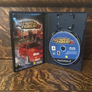 May include: Opened black PlayStation 2 game case. The cover art for "Tokyo Xtreme Racer 3" shows a red sports car racing through a city at night. The game disc is visible inside the case.