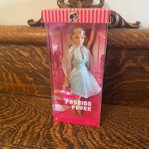 May include: A Barbie doll in a blue dress with a silver beaded bodice and a sheer overlay. The doll is packaged in a pink and white box with the words "Barbie Fashion Fever" printed on it.
