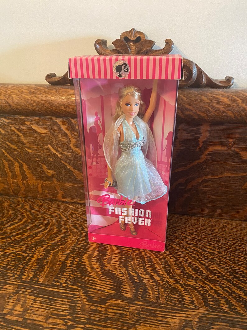 May include: A Barbie doll in a blue dress with a silver beaded bodice and a sheer overlay. The doll is packaged in a pink and white box with the words "Barbie Fashion Fever" printed on it.