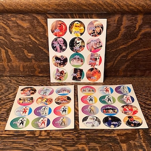 Può includere: Un set di 24 adesivi rotondi con immagini dei Power Rangers. Gli adesivi sono su tre fogli di carta bianca. Ogni adesivo ha un bordo colorato e un'immagine diversa di un Power Ranger.