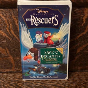 Op de afbeelding: Een VHS-band van Disney's animatiefilm "The Rescuers". De cover toont de titel in witte letters boven een illustratie van twee muizen en een pelikaan. Een groene sticker leest "Bespaar direct $2!" en de slogan luidt "Twee kleine helden. Eén groot avontuur!"