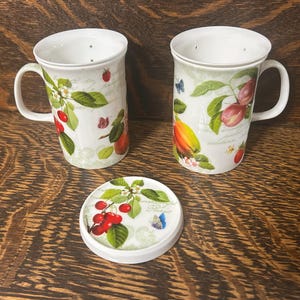 May include: Two white ceramic mugs with integrated tea strainers and a matching circular lid. The mugs feature botanical illustrations of red cherries, green leaves, and small butterflies, with faint script text printed on the white surface.