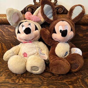 May include: Two plush Easter bunny Mickey Mouse toys. One is brown and the other is cream with a pink bow. Both have blue and white checkered ears.
