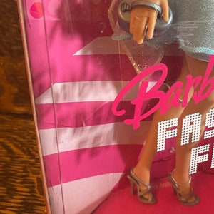 May include: A Barbie doll wearing a light blue dress with silver glitter accents. She is holding a silver clutch purse and wearing silver high heels. The doll is standing on a pink and white background with the words "Barbie Fashion Fever" printed on it.