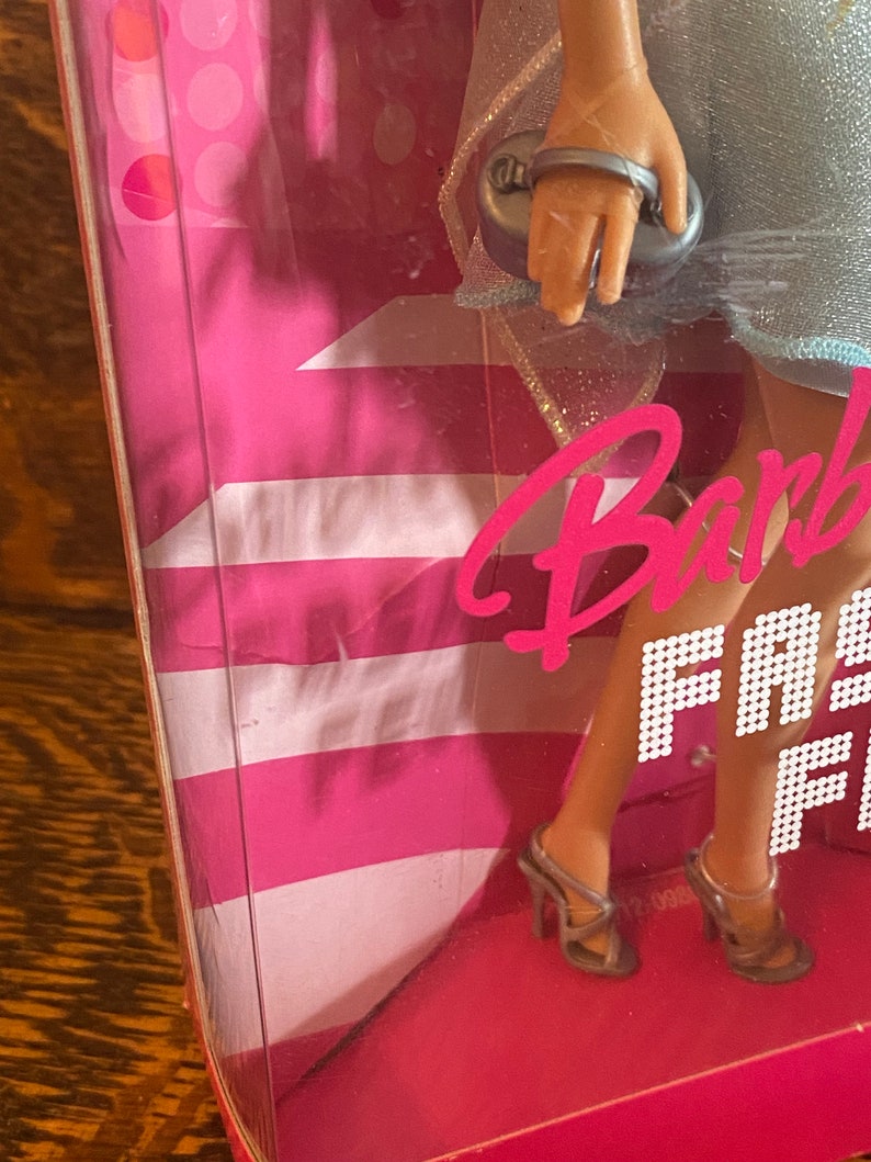 May include: A Barbie doll wearing a light blue dress with silver glitter accents. She is holding a silver clutch purse and wearing silver high heels. The doll is standing on a pink and white background with the words "Barbie Fashion Fever" printed on it.