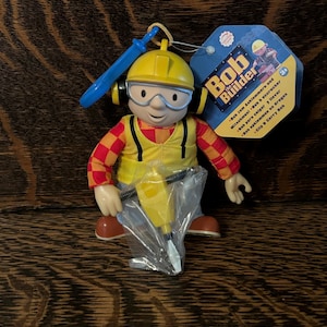 Puede incluir: Una figura de Bob el Constructor, con un casco amarillo, gafas de seguridad y un chaleco amarillo sobre una camisa a cuadros roja y naranja. La figura sostiene un martillo neumático amarillo y tiene un accesorio azul. Una etiqueta con el logo de Bob el Constructor es visible.