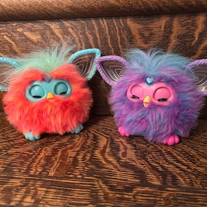 May include: Two Furby toys in vibrant colours. One is red and orange with blue details, the other is purple and blue. Both have large ears, closed eyes, and a fluffy texture. They are on a wooden surface.