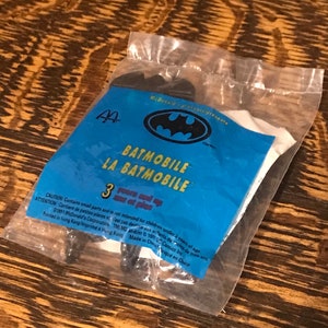 Può includere: Un sacchetto di plastica blu e giallo con un logo McDonald's e un logo Batman. Il sacchetto contiene un giocattolo Batmobile di plastica nera. Il sacchetto ha il testo "Batmobile La Batmobile" e "3 anni in su" stampato su di esso.