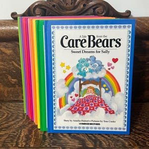 May include: A collection of colourful Care Bears children's books. The front book has a blue cover with the title "A Tale from the Care Bears: Sweet Dreams for Sally." The cover shows a rainbow, Care Bears, and a girl in bed. The books are stacked on a wooden surface.