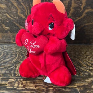 May include: A plush red devil toy with orange horns and large black eyes, holding a red heart-shaped pillow. The heart features the white embroidered text I Love You. The figure has a small red cape attached to its back and white tags attached.