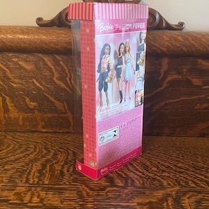 May include: A pink and white box with a Barbie logo and the words "Fashion Fever" on the front. The box contains a Barbie doll with a black and white outfit and a gold purse. The doll is standing on a pink and white background.