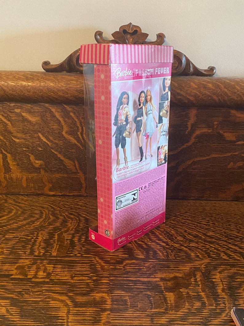 May include: A pink and white box with a Barbie logo and the words "Fashion Fever" on the front. The box contains a Barbie doll with a black and white outfit and a gold purse. The doll is standing on a pink and white background.