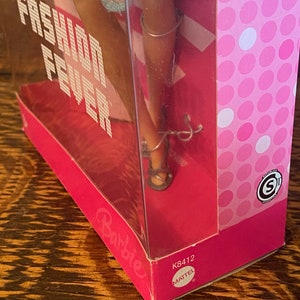 May include: A pink Barbie doll box with a clear plastic window showing a Barbie doll wearing a blue dress and silver shoes. The box has the words "Fashion Fever" printed on it. The bottom of the box has the words "Barbie" and "Mattel" printed on it.