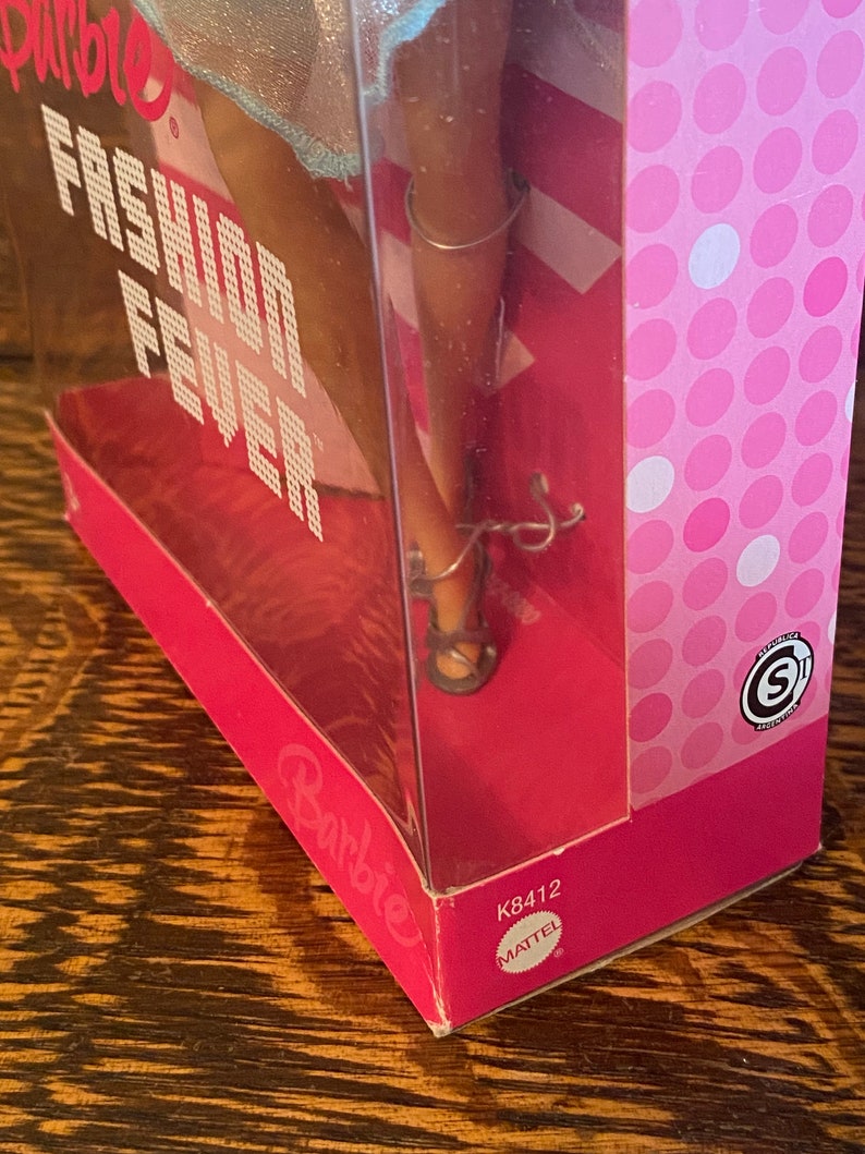 May include: A pink Barbie doll box with a clear plastic window showing a Barbie doll wearing a blue dress and silver shoes. The box has the words "Fashion Fever" printed on it. The bottom of the box has the words "Barbie" and "Mattel" printed on it.